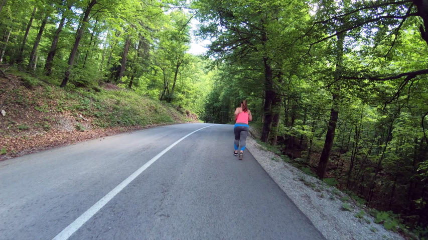 Wide forward tracking shot of an overweight woman wearing a long pony tail running on the road through a forest 