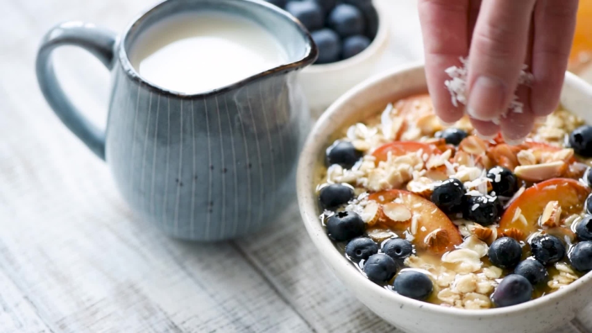 Oatmeal breakfast porridge with coconut flakes and fruits. Woman hand adding coconut flakes into porridge bowl Royalty-Free Stock Footage #1030766429