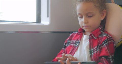 Cute Little Girl Entertaining With Tablet. Child Spending Leisure Time, Rides an Electric Train, Playing Mobile Game in the and Crushes the Bright Screen With Her Hand. Happy Family.