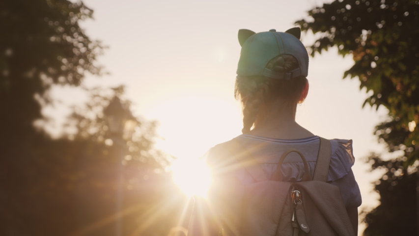 A child with a backpack is standing in the park, the setting sun shines on him