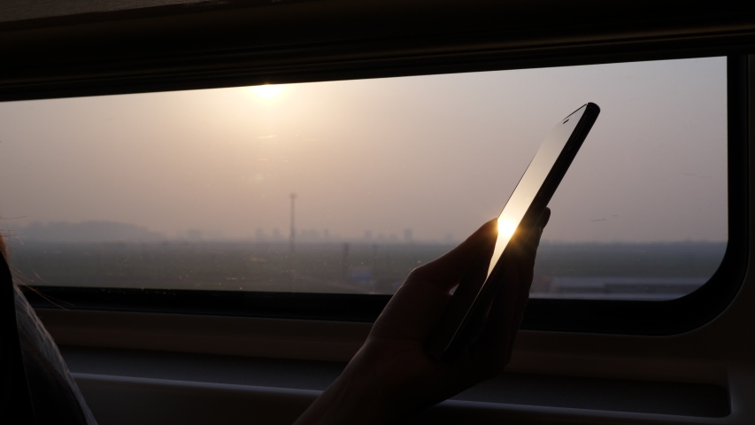 Passenger hold phone in hands, closeup high contrast shot against window, sun light shine and reflex at glossy screen of smartphone. Blurred background, modern high speed train ride to Beijing Royalty-Free Stock Footage #1030772138