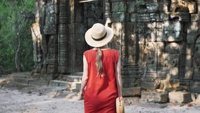 Woman in red dress and straw hat is walking among ruins of Angkor Wat temple which was built by khmer civilization in 12th century. Cambodia