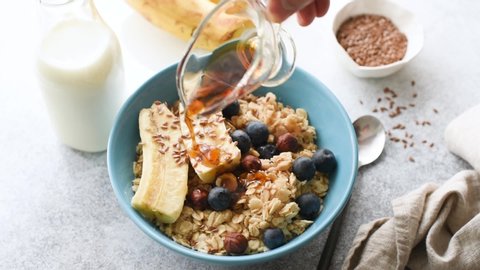 Oatmeal porridge with maple syrup, flax seeds, blueberries and banana. Person pouring maple syrup over healthy breakfast bowl