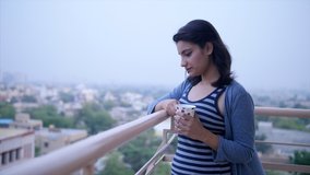 Indian girl drinking tea / coffee and looking at the view from balcony. Video of a young Asian girl standing in her house balcony and enjoying the weather - Sipping tea