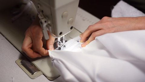 Sewing zipper on white dress. Tailor hands pulling textile under presser foot of sewing machine. Working by light of built-in hardware lamp. Cutting off thread in the end. Shooting from top angle