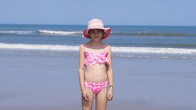 Video beautiful girls wearing in colorful hat and swimsuit enjoy playing at tropical beach. Kids enjoying warm splashing sea water on coast. Children jump on the waves on warm sunny day on seashore