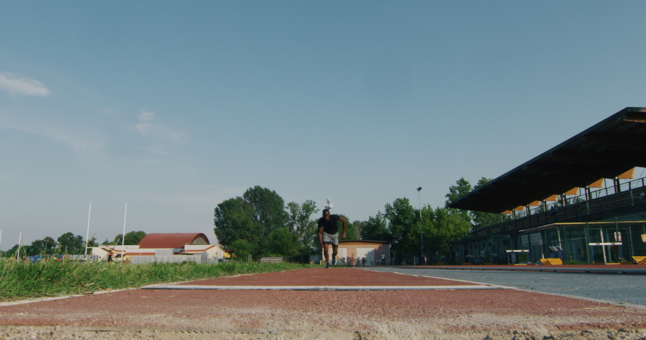 Slow motion of young african male athlete is performing a long jump on race track in athletics stadium in a sunny day.