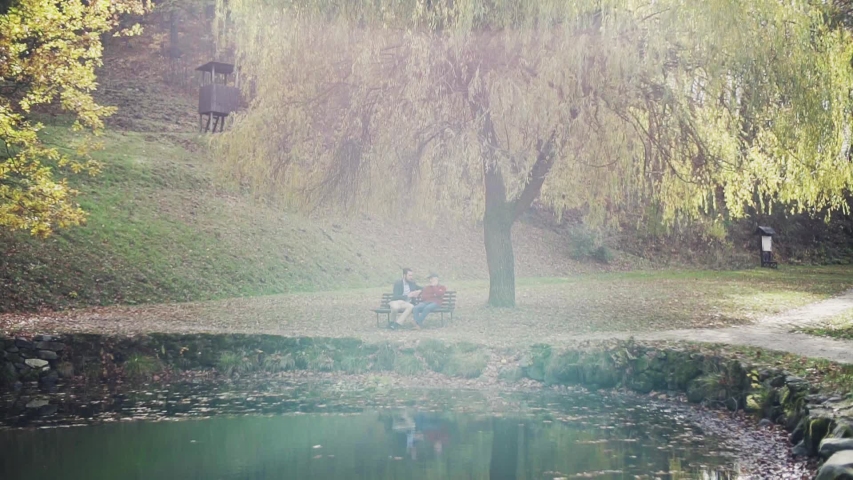 Senior father and his son sitting on bench by lake in nature, talking.