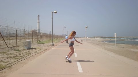 Young athletic woman surf-skating down the Tel aviv beach promenade on a bright sunny morning and turns to smile at the camera. Following shot. Reading power station in the background. - Βίντεο στοκ