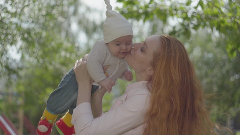 Side view of beautiful red-haired woman playing with her kid in the park or garden close-up. The lady enjoying the sunny day with her baby outdoors. Young mother throws up a child. Happy family