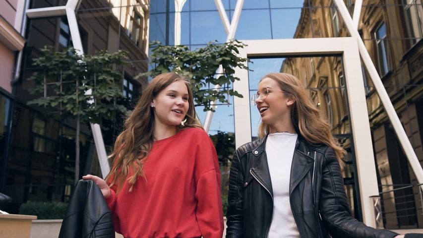 Portrait of two cheerful stylish Caucasian girlfriends walking along street near modern building while laughing and hugging each other. Outdoors, lifestyle. Slow motion
