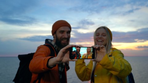 Happy, positive couple shooting video on the smartphone. Standing near the sea during the sunset.  – Stockvideo