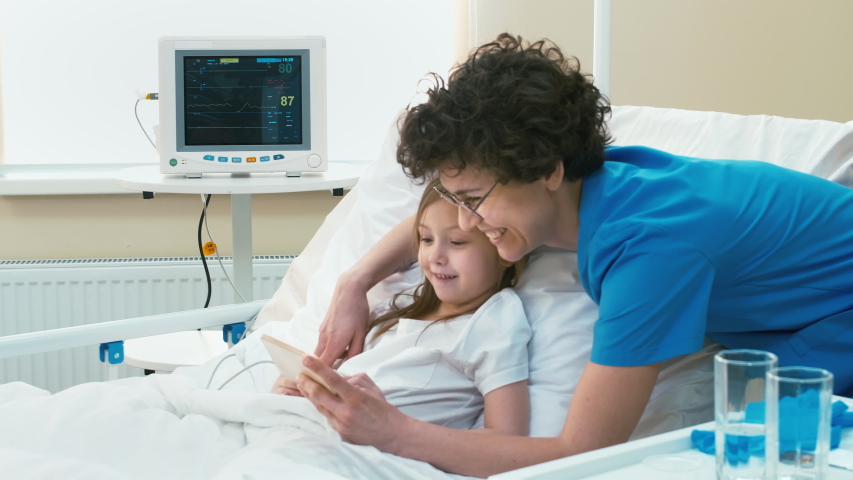 Nurse helping little patient lying in hospital bed to find cartoons in internet with mobile phone, smiling and watching it together
