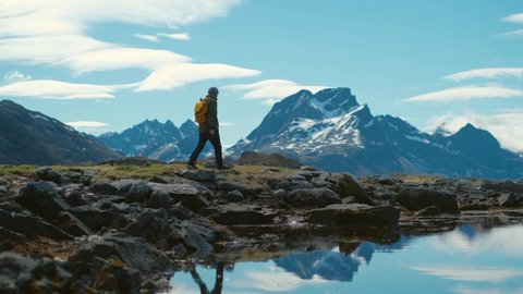 Young bearded man in a hat with a backpack comes down from the mountains on a background of snow capped mountains