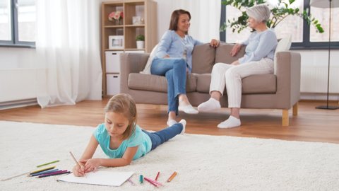 family, leisure and three generations concept - daughter drawing in sketchbook by crayons lying on floor and mother talking to grandmother on sofa at home