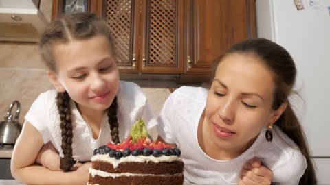 Portrat of mom and daughter are sniffing cooked homemade chocolate cream cake with blueberries, raspberries and physalis in kitchen at home. Teen girl is taking berry from the cake and eating it.