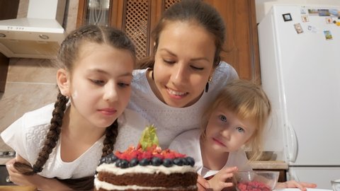 Portrat of mom and daughters are sniffing cooked homemade chocolate cream cake with blueberries, raspberries and physalis in kitchen at home. Teen girl is taking berry from the cake and eating it.