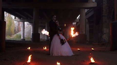 Beautiful couple in wedding attire with a terrible makeup for Halloween are standing under an abandoned bridge, in the background a huge flame of of fire show. Hellfire. Slow motion. Flamethrower.