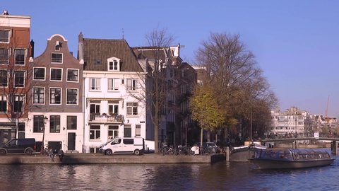 Typical canal houses in Amsterdam, The Netherlands, reflecting in the canal in front on a sunny day with blue sky following a tourist boat that comes around the corner and passes