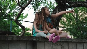 Two little girls sisters climbed onto the wall of the fence and look into the distance. Fingers show each other something interesting. Childhood concept. Outdoor video shot of child emotions in 4K.
