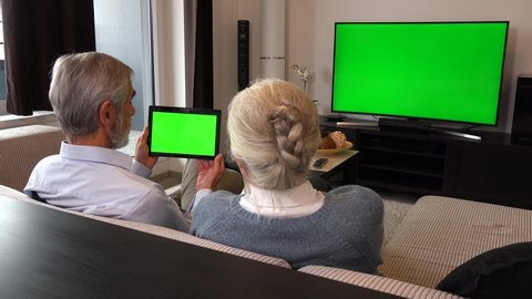 An elderly couple sits on a couch in a living room, watches TV with a green screen, looks at a tablet with a green screen and talks