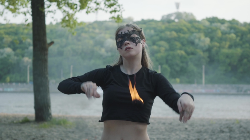 Slim woman in mask dancing, performing a show with fireball standing on the riverbank. Skillful fireshow artist showing mastery of motion of fire and graceful passionate dance