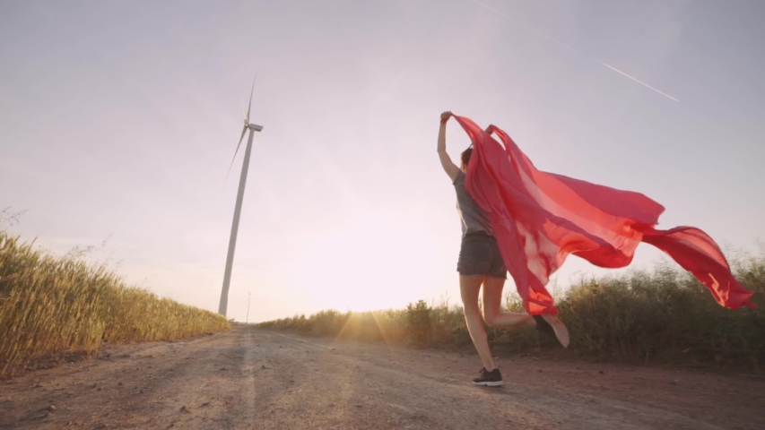 Woman with pieces of red cloth run to the wind generator in the field. Windmill on sunset. Slow motion. Stylish hipster woman