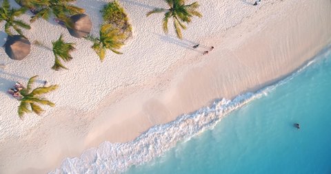 Aerial view of sea waves, umbrellas, palm trees and walking people on sandy beach at sunset. Summer in Zanzibar, Africa. Tropical landscape with parasols, sand, blue water. Top view from air. Travel