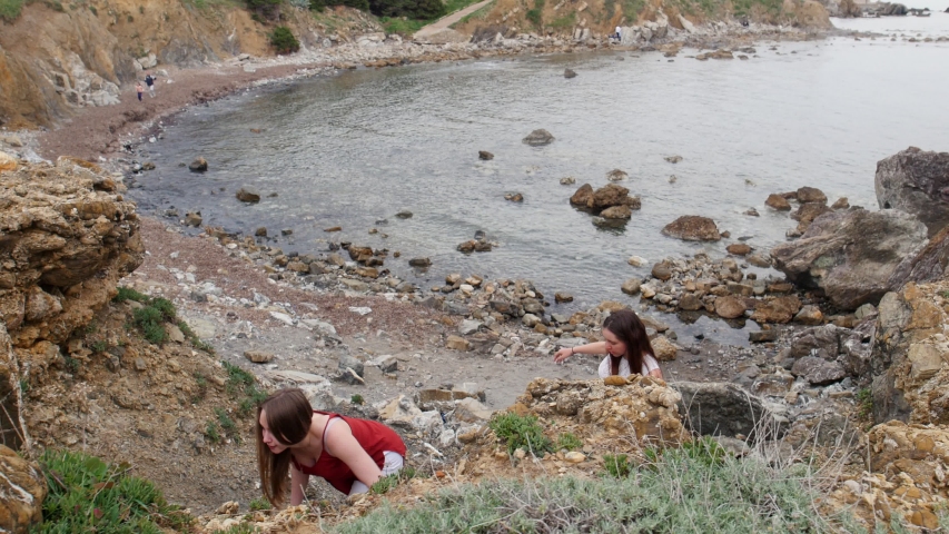 Two young women climbing up on the rocky wall from the coast