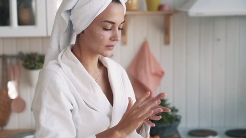 Close up of young woman in white bathrobe and with towel on head applies moisturizer cream on her face. Girl does daily beauty care cosmetic routine