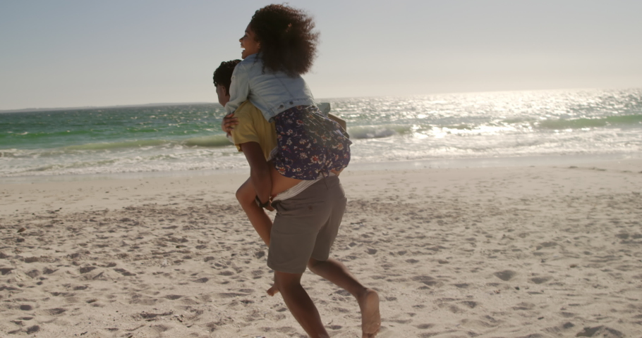 Side view of African american man giving piggyback ride to woman on the beach. They are smiling and having fun  | Shutterstock HD Video #1030933910
