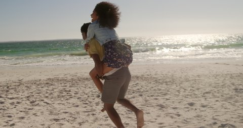 Side view of African american man giving piggyback ride to woman on the beach. They are smiling and having fun 