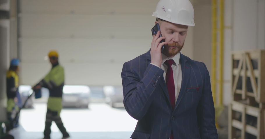 Businessman wearing hardhat speaking by phone and in workshop of modern factory. His employee works on the background | Shutterstock HD Video #1030942628