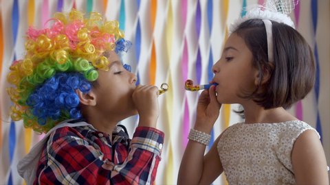 Two adorable Indian kids having fun with whistle party horns at a birthday party. A boy and a girl blowing blow horn and teasing each other - Indian birthday party celebration at home