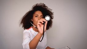 girl doctor with lush curly hair in a white robe against a white wall. in the hands of girls Stethoscope. focus transferring from the doctor's face to the stethoscope. 4K UHD video