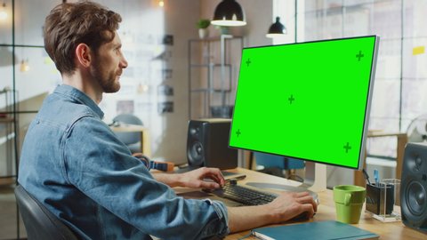 Male Video Editor with Beard Works on His Personal Computer with Big Green Screen Mock Up Display. He Works in a Cool Office Loft. Other Female Creative Colleague Walks in the Background.