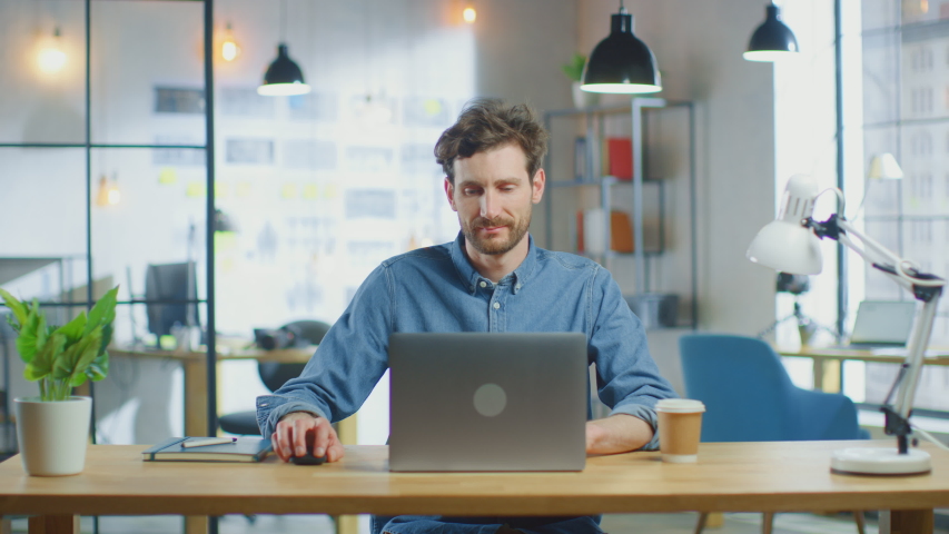 Young Handsome Man Works on a Laptop Computer in Cool Creative Agency in a Loft Office. He has a Take-away Coffee and a Notebook on the Table. He Wears a Jeans Shirt. Royalty-Free Stock Footage #1030955348