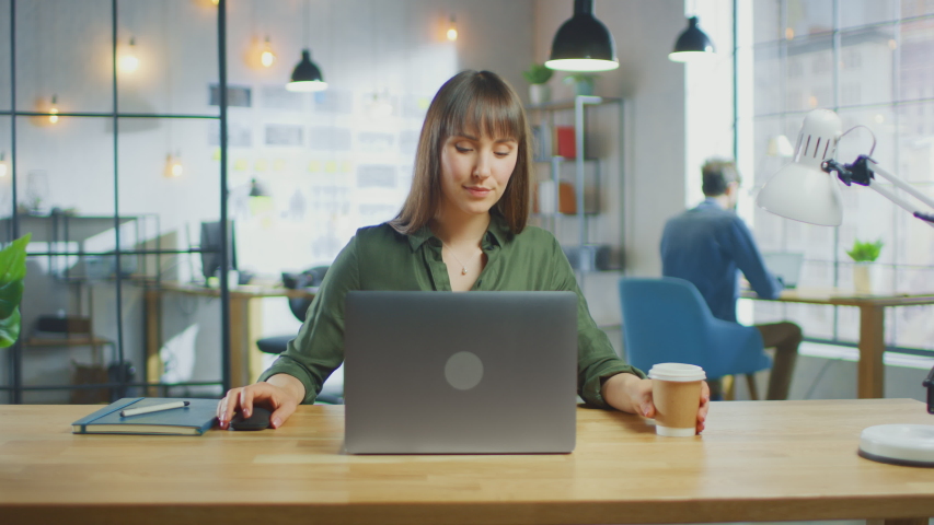 Young Beautiful Brunette Works on a Laptop Computer in Cool Creative Agency in a Loft Office. She has a Take-away Coffee and a Notebook on Her Table. Her Colleague Works in the Background. Royalty-Free Stock Footage #1030955351