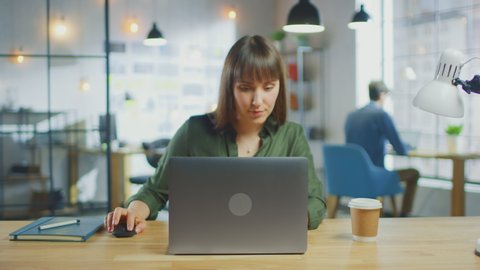Young Beautiful Brunette Works on a Laptop Computer in Cool Creative Agency in a Loft Office. She has a Take-away Coffee and a Notebook on Her Table. Her Colleague Works in the Background.