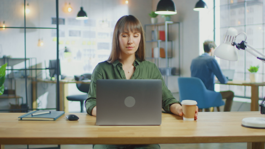 Young Beautiful Brunette Works on a Laptop Computer in Cool Creative Agency in a Loft Office. She has a Take-away Coffee and a Notebook on Her Table. Her Colleague Works in the Background. Royalty-Free Stock Footage #1030955354