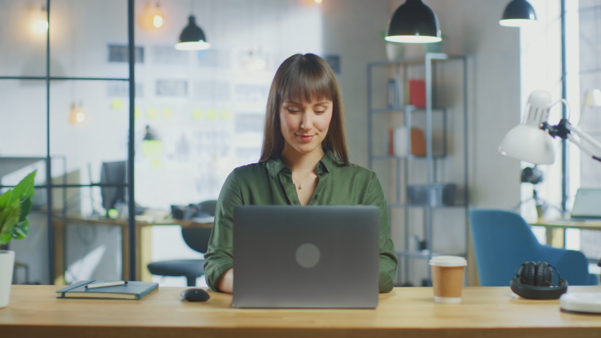 Young Beautiful Brunette Works on a Laptop Computer in Cool Creative Agency in a Loft Office. She has a Take-away Coffee and a Notebook on Her Table. Camera Zooms In and She Smiles and Laughs. | Shutterstock HD Video #1030955399