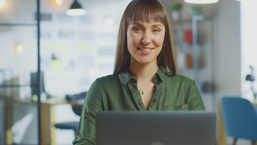 Young Beautiful Brunette Works on a Laptop Computer in Cool Creative Agency in a Loft Office. She has a Take-away Coffee and a Notebook on Her Table. Camera Zooms In and She Smiles and Laughs. | Shutterstock HD Video #1030955399