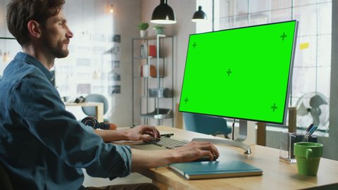 Young Professional Creative Employee Sits Down and Works on His Personal Computer with Big Green Screen Mock Up Display. He Works in a Cool Office Loft. Other Female Colleague Walks in the Background.
