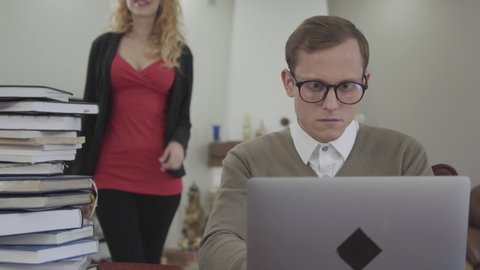Young modestly dressed man in glasses sitting at table at home, working on laptop. Beautiful woman with deep neckline bends over and putting hand on his shoulder. The nerd trying to shake off the hand