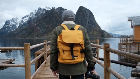 Traveler goes on a wooden pier. dressed in a jacket and knitted hat and a yellow backpack. back view. Norway lofoten