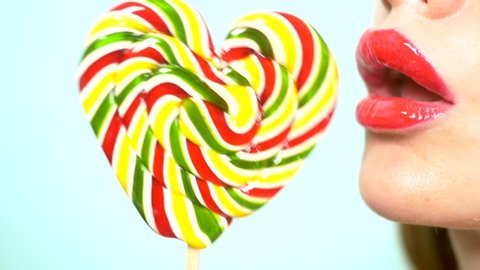 close-up, female lips with red lipstick, eat and lick a big lollipop in the shape of a heart. copy space, blue background