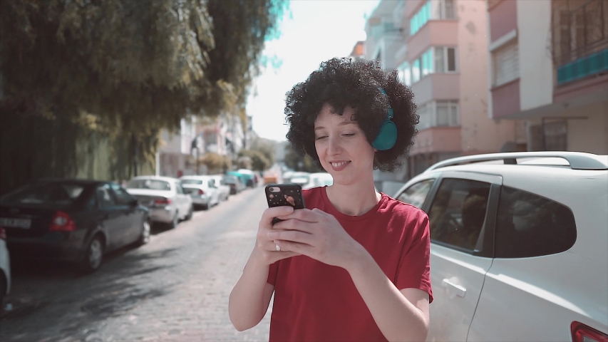 Afro haired young girl is walking when she is using mobile phone and taking selfie. listening music with headphone. Texting, using smartphone. 