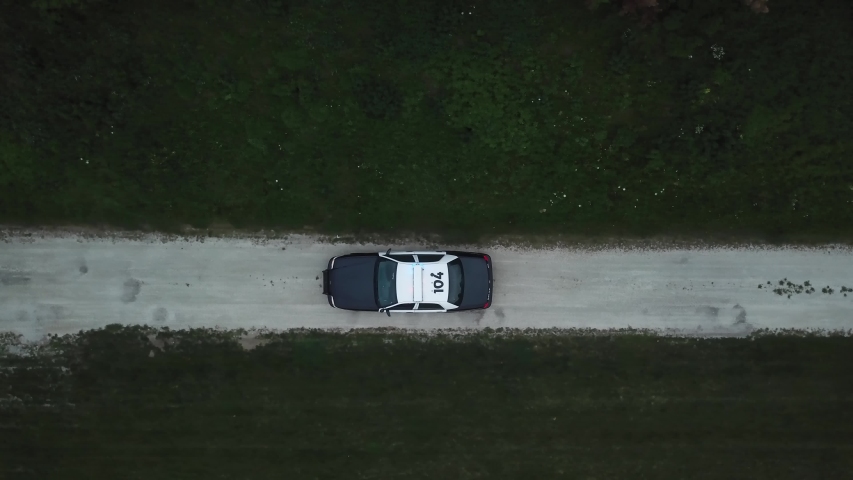 AERIAL: angled down track of a police car traveling through a dirt road with its lights flashing.