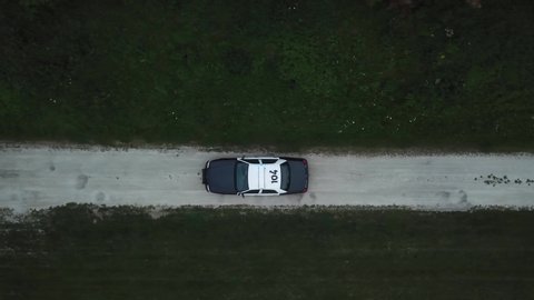 AERIAL: angled down track of a police car traveling through a dirt road with its lights flashing.