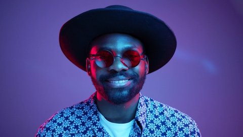 Cheerful stylish Afro American hipster man in trendy hat and stylish steampunk polarized eyeglasses attractively smiling straight to camera on bright colorful neon lights. Studio portrait. Style, new